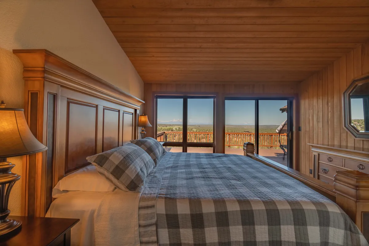A tranquil bedroom featuring a plaid bedspread, wooden headboard, and large windows with an inviting view outdoors. The room exudes restful charm and privacy.