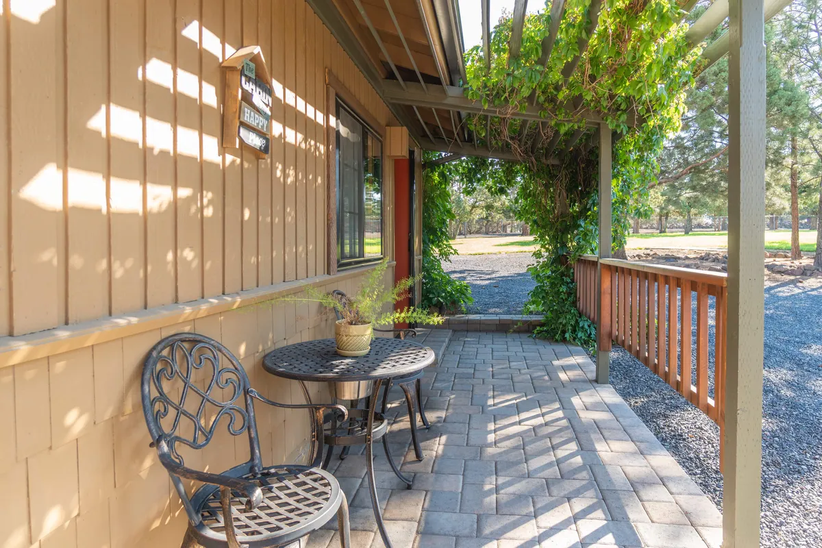 A cozy exterior patio scene with a table and chairs surrounded by lush planters and vines climbing a trellis. The wooden railing and shaded setting create an inviting outdoor space ideal for gathering, relaxing, and taking in the peaceful surroundings.