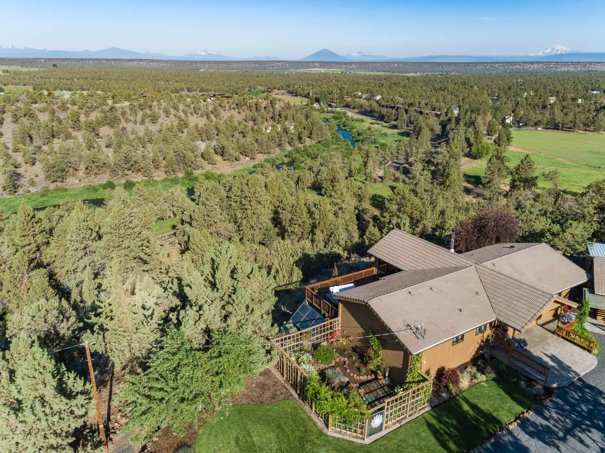The exterior shows the house nestled among trees, bordered by a lush garden and distant mountains. The scene evokes a tranquil, natural setting with opportunities for gardening, yard games, and enjoying wide open spaces.