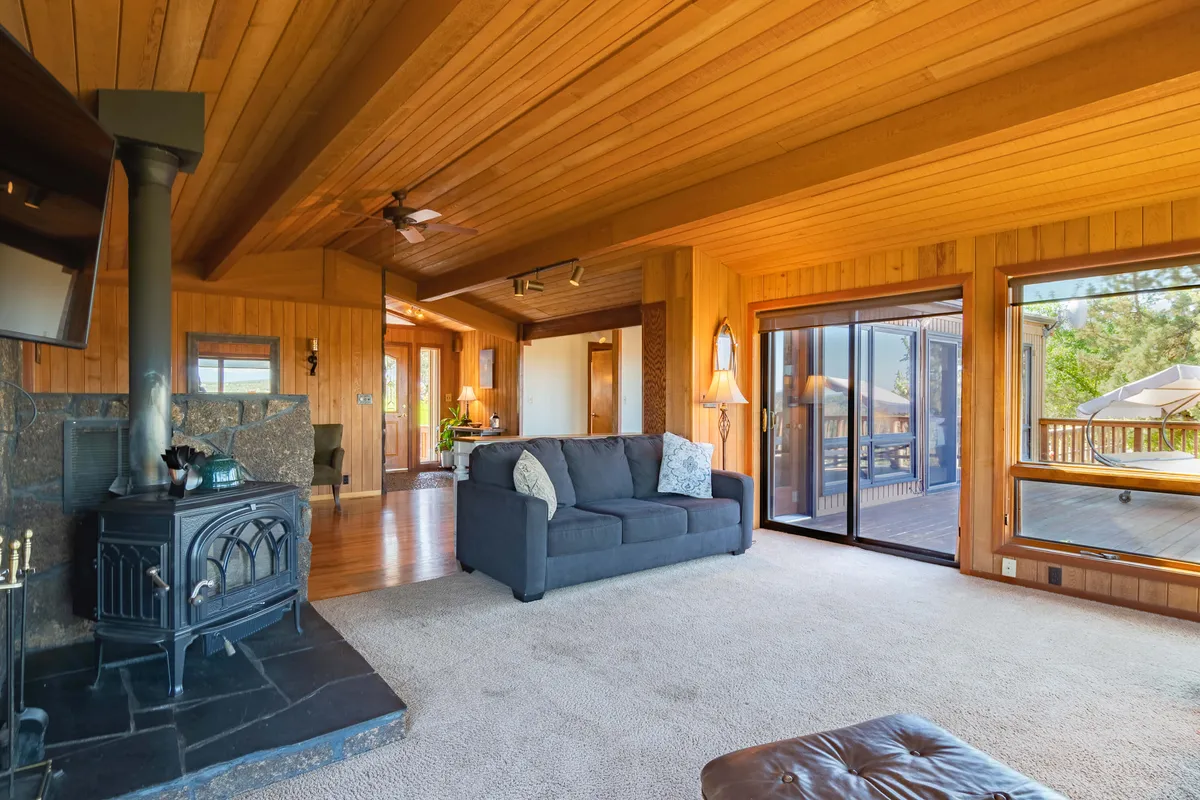 This image captures a warm, inviting living room with a wooden ceiling, a cozy fireplace, plush sofas, and expansive windows that frame stunning views of the mountains and river. The ambiance is perfect for relaxing, socializing, or enjoying quiet moments while soaking in the natural landscape.